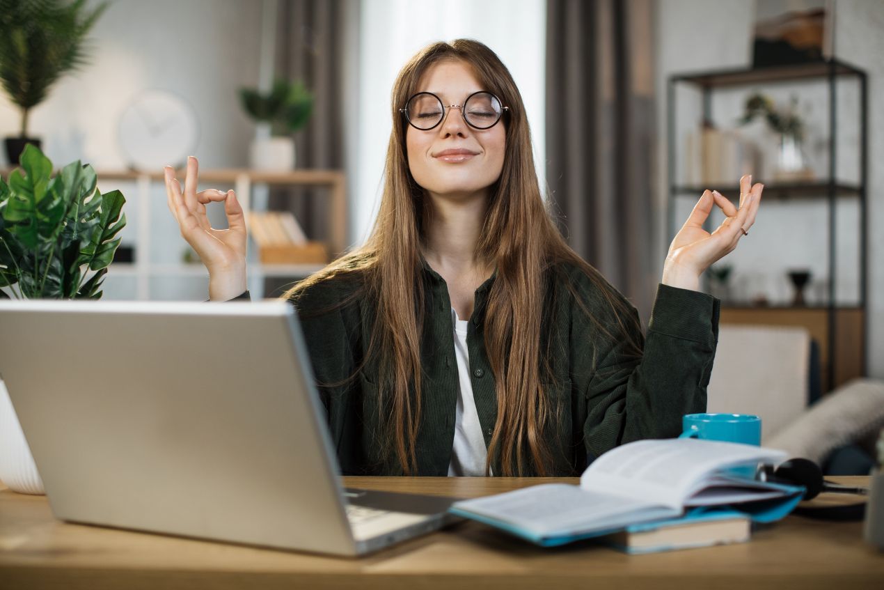 Eine junge Frau meditiert am Laptop zu Hause, Symbol fuer Konfliktloesung am Arbeitsplatz durch Achtsamkeit und Stressabbau