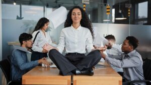Eine Frau sitzt meditierend auf einem Schreibtisch, waehrend Kollegen streiten – Symbol fuer Konfliktloesung am Arbeitsplatz und mentale Balance im Job